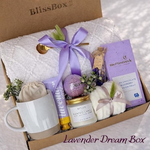 May include: A gift box labeled BlissBox containing a white textured blanket tied with a purple ribbon, a Moonstruck dark chocolate bar, a lavender bath bomb, a white ceramic mug, a lavender sage tube, a scented candle, and a small white pouch.