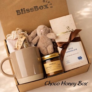 May include: A gift box labeled BlissBox containing a beige ceramic mug, a small plush brown rabbit, a glass jar with dried flowers, a vanilla scented candle in a glass jar, and a Moonstruck milk chocolate bar tied with a brown ribbon.