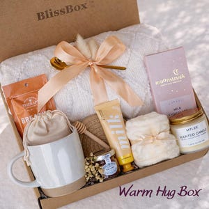 May include: A gift box containing a white cable-knit blanket tied with a peach ribbon, a ceramic mug, a pouch, a honey dipper, a jar of honey, a tube of milk lotion, a bar of Moonstruck milk chocolate, and a scented candle by Mtlee.