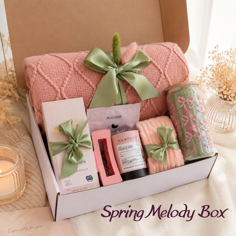 May include: A gift box containing a pink textured knit blanket tied with a sage green ribbon, a chocolate bar, a Peauamie face mask, a pink tea infuser, a Tea With Tae tea tin, a pair of pink socks, and a glass tumbler with pink bow patterns.