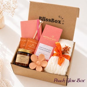 May include: A cardboard gift box labelled BlissBox containing a coral notebook with a gold pen, two Harney & Sons tea packets, a Moonstruck dark chocolate bar, a Bergamot candle, a peach-coloured flower hair clip, and a white fuzzy item with an orange ribbon.