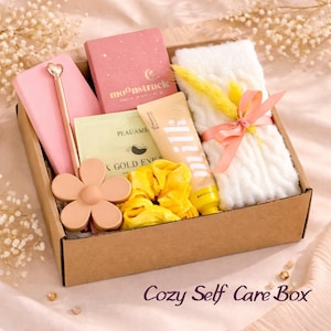 May include: A brown cardboard box containing self-care products. It features a pink notebook and pen, a pink flower-shaped object, a pink box, a yellow scrunchie, a lotion tube, a face mask, and a white knitted item tied with a pink ribbon. The text "Cozy Self Care Box" is at the bottom.