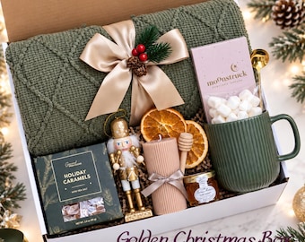 May include: A gift box containing a folded olive green knit blanket with a champagne ribbon, a Moonstruck chocolate bar, a green ribbed mug with a gold spoon, a nutcracker, a pillar candle, dried orange slices, honey, and holiday caramels.