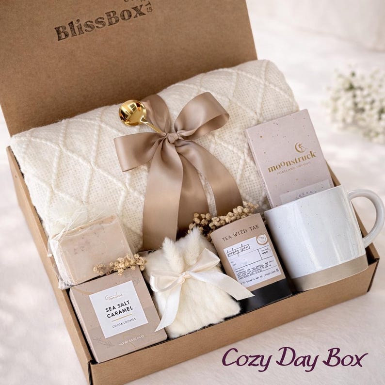 Op de afbeelding: Een BlissBox geschenkdoos met een cr&egrave;mekleurige gebreide deken vastgebonden met een gouden lint en een gouden lepel, een Moonstruck chocoladereep, een witte keramische mok, een Tea With Tae theeblik, een doosje Sea Salt Caramel koekjes en een bontzakje.