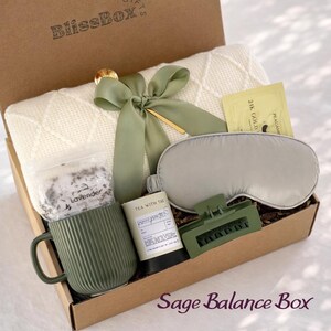 May include: A gift box labeled "BlissBox" filled with self-care items. Includes a cream-colored blanket, a sage green mug, a sleep mask, a hair clip, tea, bath salts, and a gold-accented bow. The box is labeled "Sage Balance Box".