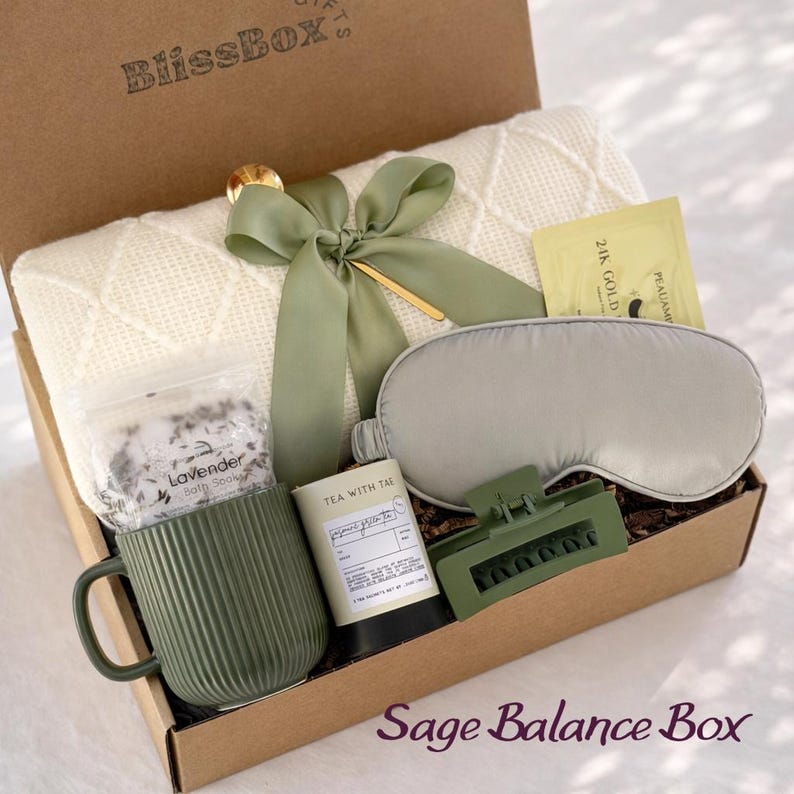 May include: A gift box labeled "BlissBox" filled with self-care items. Includes a cream-colored blanket, a sage green mug, a sleep mask, a hair clip, tea, bath salts, and a gold-accented bow. The box is labeled "Sage Balance Box".