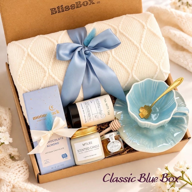 Op de afbeelding: Een geschenkdoos met een cr&egrave;mekleurige kabelgebreide deken vastgebonden met een blauw lint, een lichtblauwe chocoladereep, een theeblik, een geurkaars, een klein potje honing met een houten lepeltje en een lichtblauw theekopje met een gouden lepel.