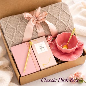Op de afbeelding: Een geschenkdoos met een beige, getextureerde plaid vastgebonden met een roze satijnen lint, een roze notitieboek met een gouden pen met hartvormige top, een doosje met Hot Cocoa Velvety White en een roze geschulpt theekopje met schotel en gouden lepel.