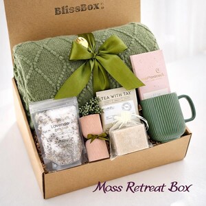 May include: A gift box labeled "BlissBox" filled with self-care items. It includes a green blanket, a green mug, a pink box, a bag of lavender bath soak, a bar of soap, and a tea bag. The box is tied with a green ribbon and the words "Mass Retreat Box" are written on the bottom.