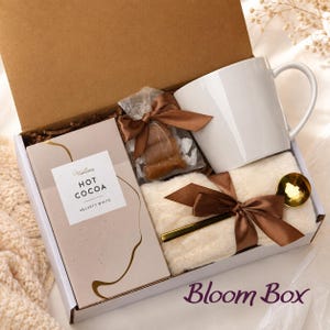 Op de afbeelding: Een geschenkdoos met een witte keramische mok, een doos Gracious Hot Cocoa Velvety White, een klein doorzichtig zakje met bruine snoepjes met een bruin lint, een gevouwen cr&egrave;mekleurig textiel en een goudkleurige lepel, alles samengebonden met lint.