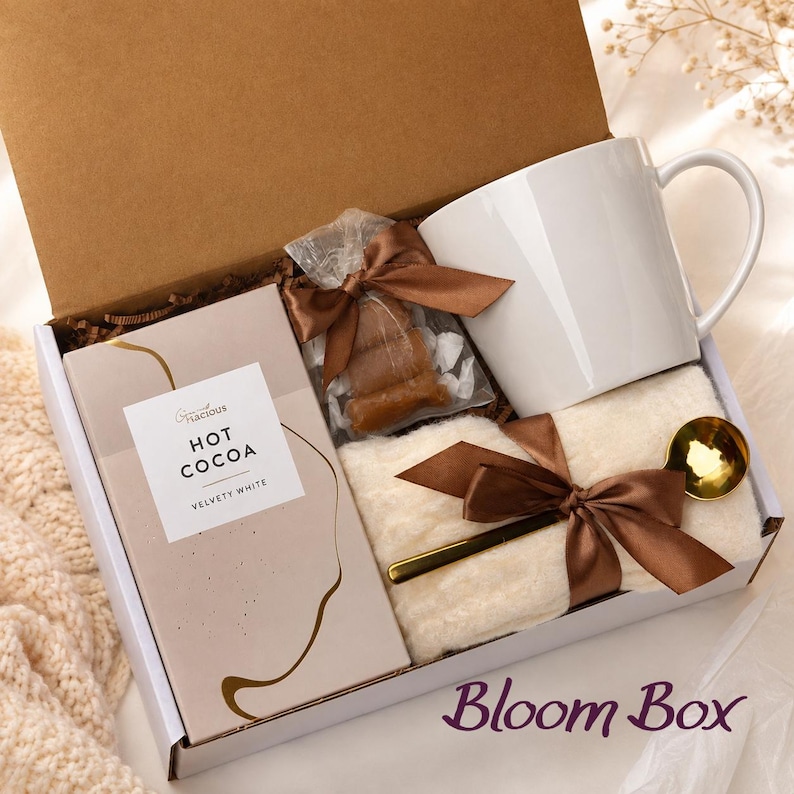 Op de afbeelding: Een geschenkdoos met een witte keramische mok, een doos Gracious Hot Cocoa Velvety White, een klein doorzichtig zakje met bruine snoepjes met een bruin lint, een gevouwen cr&egrave;mekleurig textiel en een goudkleurige lepel, alles samengebonden met lint.