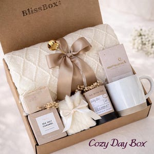 May include: A gift box labeled BlissBox containing a cream-colored knit blanket tied with a gold ribbon and a gold spoon, a Moonstruck chocolate bar, a white ceramic mug, a Tea With Tae tea tin, a Sea Salt Caramel cookie box, and a white fur pouch.
