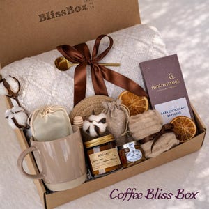 May include: A gift box labeled BlissBox containing a white textured throw blanket tied with a brown ribbon, a beige ceramic mug, a jar of Bergamot candle, a small jar of honey, a Moonstruck dark chocolate espresso bar, cotton stems, and dried orange slices.