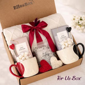 May include: A gift box containing a white knit blanket tied with a red ribbon, two mugs with heart-shaped designs, two packets of hot cocoa with marshmallows, a bag of lavender bath salts, and two small heart-shaped ornaments in red and white.
