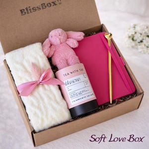 Puede incluir: Una caja de regalo con un conejito de peluche rosa, un cuaderno rosa, un bol&iacute;grafo dorado, una manta blanca atada con una cinta rosa y un bote de t&eacute;. El bote de t&eacute; dice "Tea with Tae" y "Hibiscus Sage". La caja est&aacute; etiquetada como "BlissBox" y "Soft Love Box".