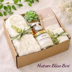 May include: A gift box labeled "Nature Bliss Box" filled with self-care items. The box contains a white fluffy pair of mittens, a small succulent, a jar of honey, a scented candle, and a small notebook. The items are tied with green ribbons.
