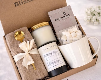 May include: A brown BlissBox gift set with a candle, tea, mug, spoon, and a soft blanket tied with a bow. The box includes a tea packet from Moonstruck and a jar of marshmallows. The words "Tea with Tae" are visible on a tea container.