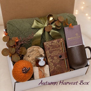 May include: A gift box containing a folded olive green quilted blanket tied with a satin ribbon, a brown ribbed mug, a brown notebook with gold leaf patterns and a pen, a small jar of honey, a knitted orange pumpkin, and a chocolate bar.