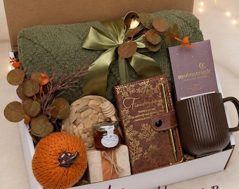 May include: A gift box containing a folded olive green quilted blanket tied with a satin ribbon, a brown ribbed mug, a brown notebook with gold leaf patterns and a pen, a small jar of honey, a knitted orange pumpkin, and a chocolate bar.
