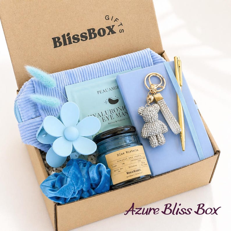 May include: A cardboard gift box labeled BlissBox containing a light blue corduroy pouch, a flower-shaped hair clip, a blue velvet scrunchie, a hyaluronic eye mask, a blue candle, a light blue notebook, a gold pen, and a rhinestone bear keychain.