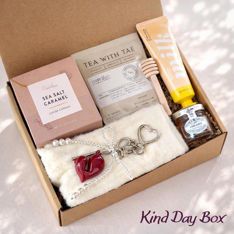 May include: A gift box filled with various items, including a box of Sea Salt Caramel Cocoa Cookies, tea bags, a tube of milk lotion, a small jar of honey, and a heart-shaped keychain. The box is lined with a fluffy white material. The text "Kind Day Box" is visible.