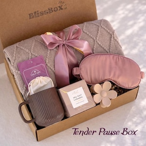 Op de afbeelding: Een geschenkdoos met een beige kabelgebreide deken vastgebonden met een mauve satijnen lint, een mauve slaapmasker, een bruine geribbelde keramische mok, een klein wit trekkoordzakje, een bloemvormige haarklem en een doosje met Sea Salt Caramel.