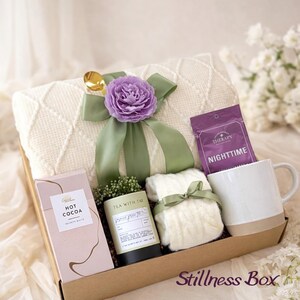 May include: A gift box with a cream-colored blanket, a purple flower, and a green ribbon. The box also contains a mug, hot cocoa, tea, socks, and a packet of nighttime tea. The words "Stillness Box" are printed on the box.