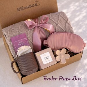 May include: A gift box labeled "BlissBox" contains a gray knit blanket, a pink satin sleep mask, a brown mug, a tea packet, a box of sea salt caramel cookies, a flower-shaped hair clip, and a pink ribbon. The box is labeled "Tender Pause Box".