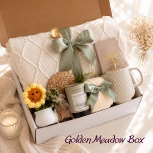 May include: A gift box containing a cream-coloured cable knit blanket tied with a sage green ribbon, a ribbed ceramic mug, a candle labelled Tea with Tale, a small crocheted sunflower in a white pot, a woven coaster, and a gold-toned spoon.