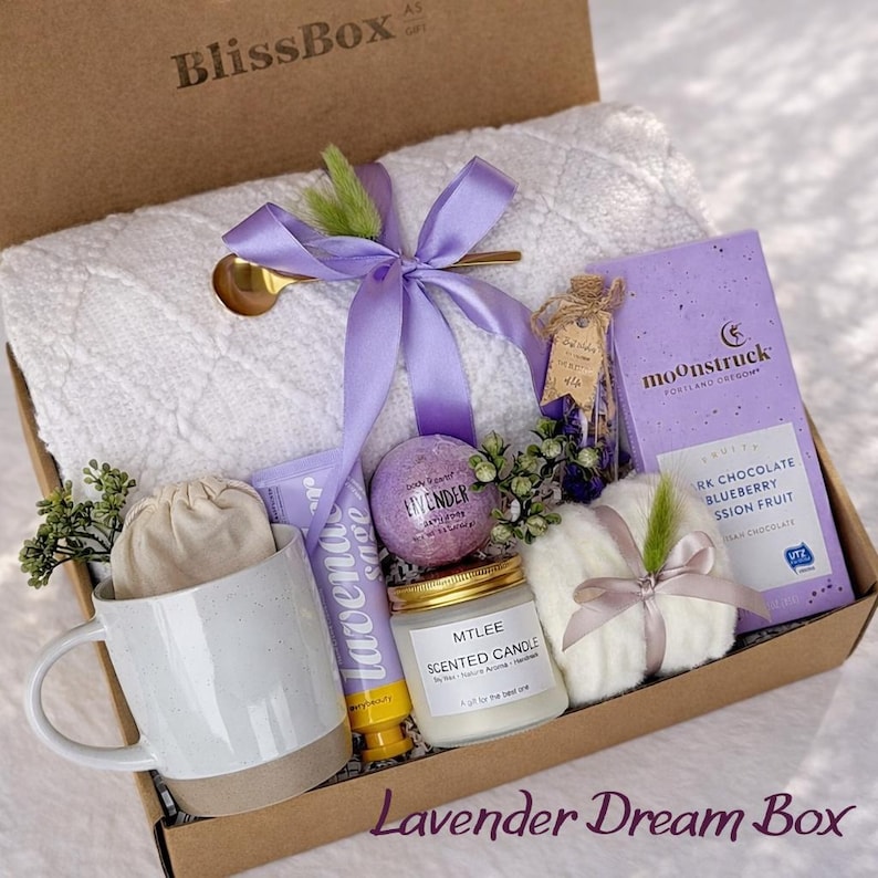 May include: A gift box filled with self-care items. Includes a white towel, a mug, a candle, a bath bomb, a chocolate bar, and other lavender-scented products. The box is tied with a purple ribbon and has the text "Lavender Dream Box".