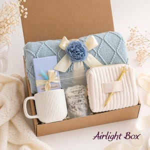 May include: A gift box containing a light blue diamond-patterned knit blanket with a blue flower and cream ribbon, a white ribbed ceramic mug, a chocolate bar with a ribbon, a pouch of lavender bath salts, a cream corduroy pouch, and a gold pen.