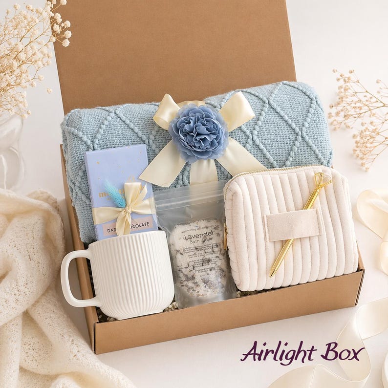 May include: A gift box containing a light blue diamond-patterned knit blanket with a blue flower and cream ribbon, a white ribbed ceramic mug, a chocolate bar with a ribbon, a pouch of lavender bath salts, a cream corduroy pouch, and a gold pen.