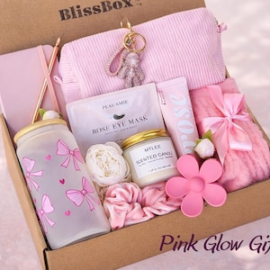 May include: A gift box labelled "BlissBox" containing pink items. Includes a pink journal, pen, cosmetic bag, eye mask, candle, tumbler, and a pink flower. The box is decorated with pink bows and hearts. Text reads "Pink Glow Gift".