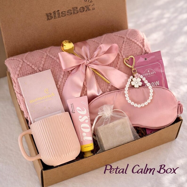 May include: A pink gift box labeled "BlissBox" filled with self-care items. Includes a pink blanket, mug, eye mask, hand cream, soap, and a gold spoon. A pink ribbon and a pearl and flower keychain add decorative touches. The box is labeled "Petal Calm Box".