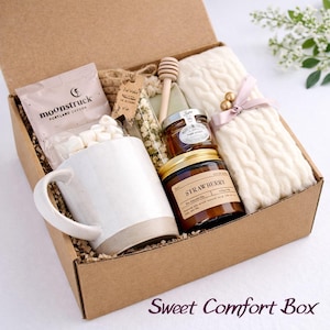 May include: A gift box filled with comfort items. Includes a white mug, a packet of marshmallows, a jar of honey, a strawberry-scented candle, a wooden honey dipper, and a cream-colored knitted scarf. The box is labeled "Sweet Comfort Box".