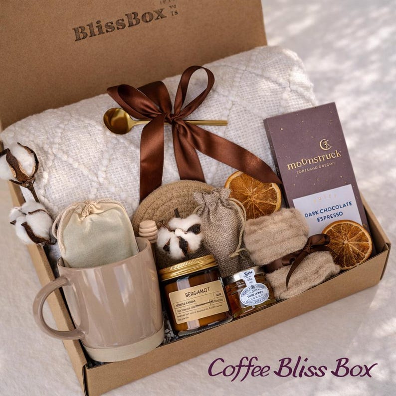 May include: A brown cardboard gift box labeled "BlissBox" contains a white blanket, a mug, a candle, a chocolate bar, and other items. The box is tied with a brown ribbon and includes the text "Coffee Bliss Box".
