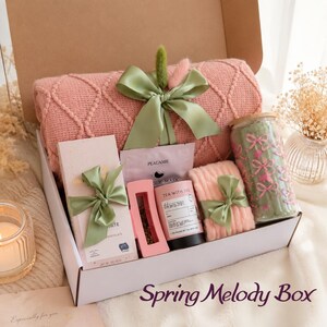 May include: A gift box labeled "Spring Melody Box" filled with a pink knitted blanket, chocolate, tea, a face mask, and a jar of green-colored items. Each item is tied with a green ribbon.