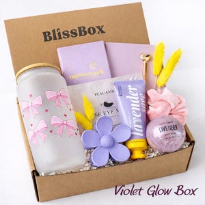 May include: A BlissBox gift set containing a frosted glass tumbler with pink bows, a lavender hand cream tube, a purple flower hair clip, a lavender bath bomb, a Moonstruck notebook, a gold pen, yellow dried grass, and a pink scrunchie.