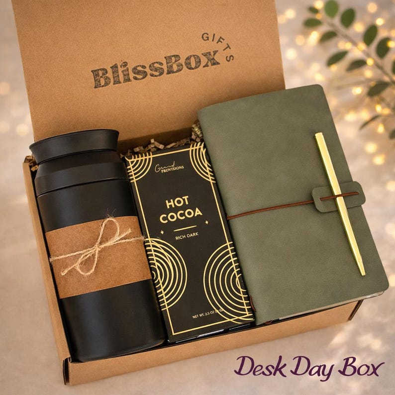 May include: A gift box labeled "BlissBox Gifts" contains a black travel mug with a brown paper wrap, a dark chocolate bar labeled "Hot Cocoa", and a green notebook with a gold pen holder. The box is labeled "Desk Day Box".