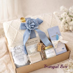 Op de afbeelding: Een geschenkdoos met een witte, getextureerde deken vastgebonden met een blauw lint en bloem, een zakje lavendelbadzout, een doosje Tea with Tae, een stuk zeep, een glazen buisje met gedroogde bloemen en een Moonstruck chocoladereep.
