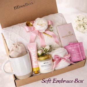 May include: A gift box labeled BlissBox containing a white cable knit blanket, a white ceramic mug, a beige drawstring pouch, a rose hand cream tube, a scented candle, a pink ribbon, a gold spoon, a Moonstruck chocolate bar, and raspberry tea.