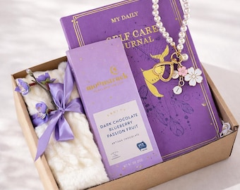 May include: A gift box containing a purple journal with the text "MY DAILY SELF CARE JOURNAL", a bar of dark chocolate, a white scarf, and a pearl charm. The chocolate bar reads "DARK CHOCOLATE BLUEBERRY PASSION FRUIT". The box is tied with a purple ribbon.