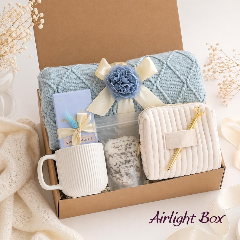 May include: A gift box containing a light blue diamond-patterned knit blanket with a blue flower and cream ribbon, a white ribbed ceramic mug, a chocolate bar with a ribbon, a pouch of lavender bath salts, a cream corduroy pouch, and a gold pen.