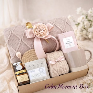 Op de afbeelding: Een geschenkdoos met een mauve gebreide deken met lint en bloem, een doos warme chocolademelk, een beige mok, een paar kabelgebreide sokken, een theezakje met het label Tea With Tae, een honingdipper, een pot honing en een kaars met het label Gardenia.