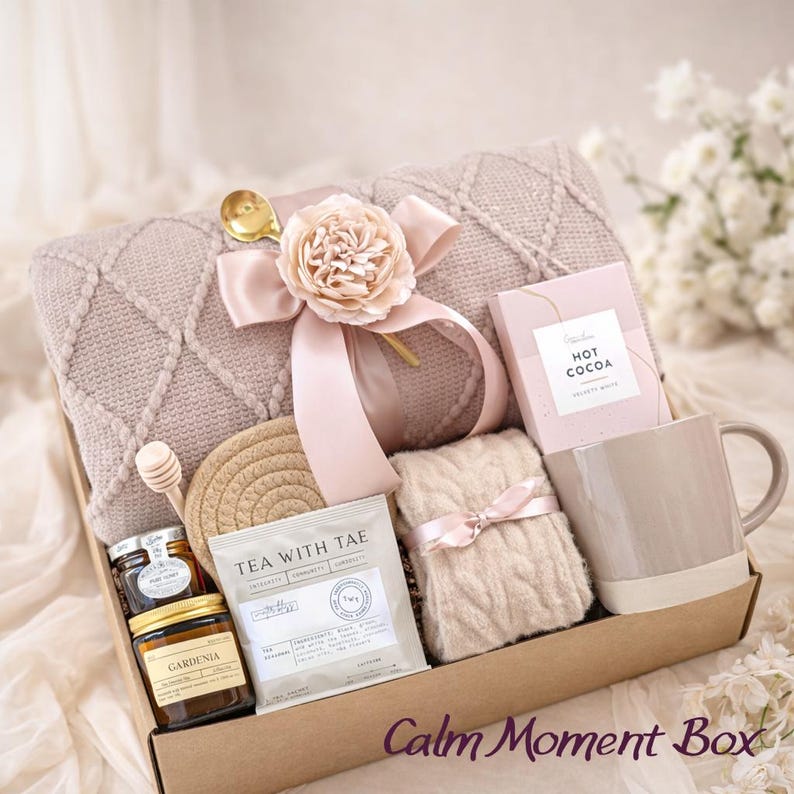 Op de afbeelding: Een geschenkdoos met een mauve gebreide deken met lint en bloem, een doos warme chocolademelk, een beige mok, een paar kabelgebreide sokken, een theezakje met het label Tea With Tae, een honingdipper, een pot honing en een kaars met het label Gardenia.