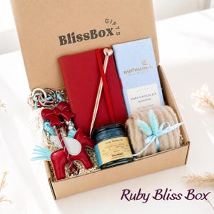 May include: A gift box labeled "BlissBox" contains a red notebook, a pen with a heart-shaped top, a blue candle, a bar of dark chocolate, a beige scarf tied with a blue ribbon, and a red horse keychain. The box is labeled "Ruby Bliss Box".