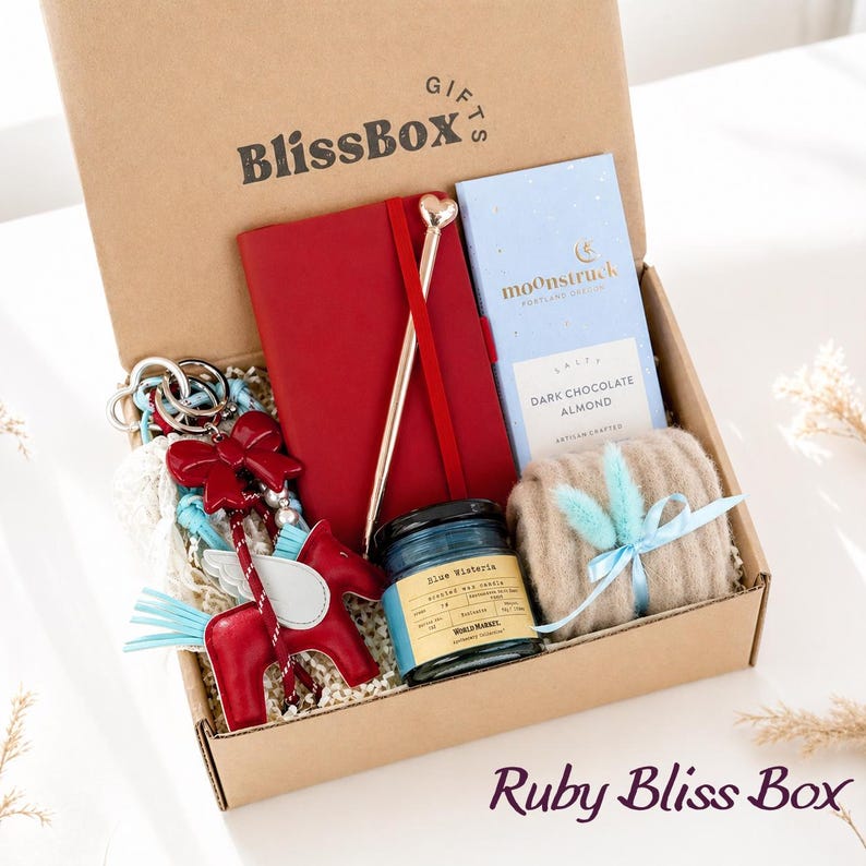 May include: A gift box labeled "BlissBox" contains a red notebook, a pen with a heart-shaped top, a blue candle, a bar of dark chocolate, a beige scarf tied with a blue ribbon, and a red horse keychain. The box is labeled "Ruby Bliss Box".