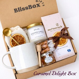 May include: A cardboard gift box labeled BlissBox containing a white ceramic mug, a gold spoon, a scented candle in a glass jar, a wooden honey dipper, a small jar of honey, individually wrapped caramels, and a Moonstruck chocolate bar with a dried orange slice.