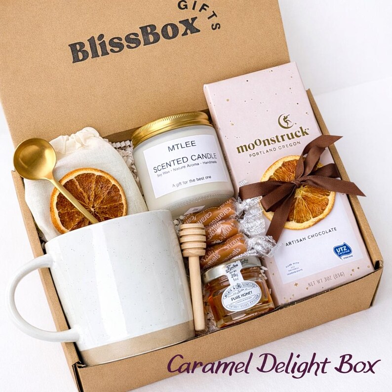May include: A cardboard gift box labeled BlissBox containing a white ceramic mug, a gold spoon, a scented candle in a glass jar, a wooden honey dipper, a small jar of honey, individually wrapped caramels, and a Moonstruck chocolate bar with a dried orange slice.