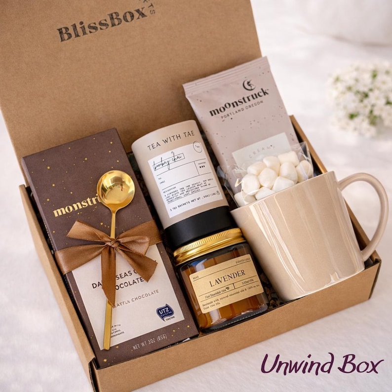Puede incluir: Una caja de regalo de cart&oacute;n marr&oacute;n etiquetada como "BlissBox" llena de art&iacute;culos de relajaci&oacute;n. Incluye una barra de chocolate, cuchara dorada, t&eacute;, tarro de lavanda, malvaviscos y una taza beige. La caja est&aacute; etiquetada como "Unwind Box".